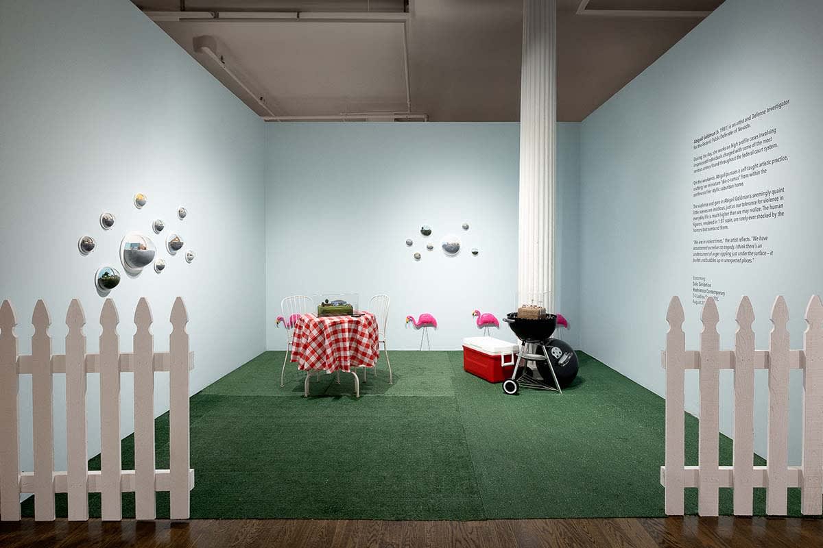 Abigail Goldman installation at Outsider Art Fair. Picturesque suburban backyard with white picket fence in forground, red checkered tablecloth bistro table and grill / cooler visible. Abigail's work hangs in clusters on walls and on top of grill and tabl