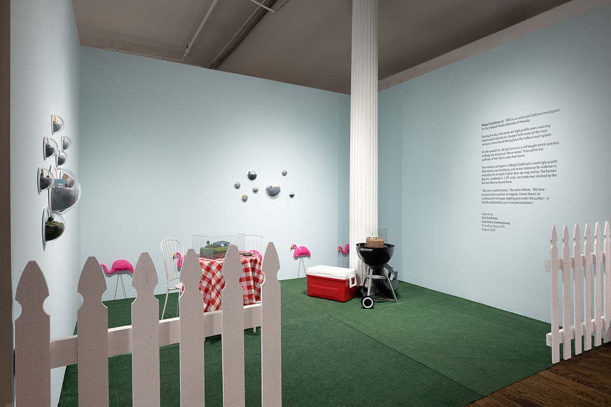Abigail Goldman installation at Outsider Art Fair. Picturesque suburban backyard with white picket fence in forground, red checkered tablecloth bistro table and grill / cooler visible. Abigail's work hangs in clusters on walls and on top of grill and tabl