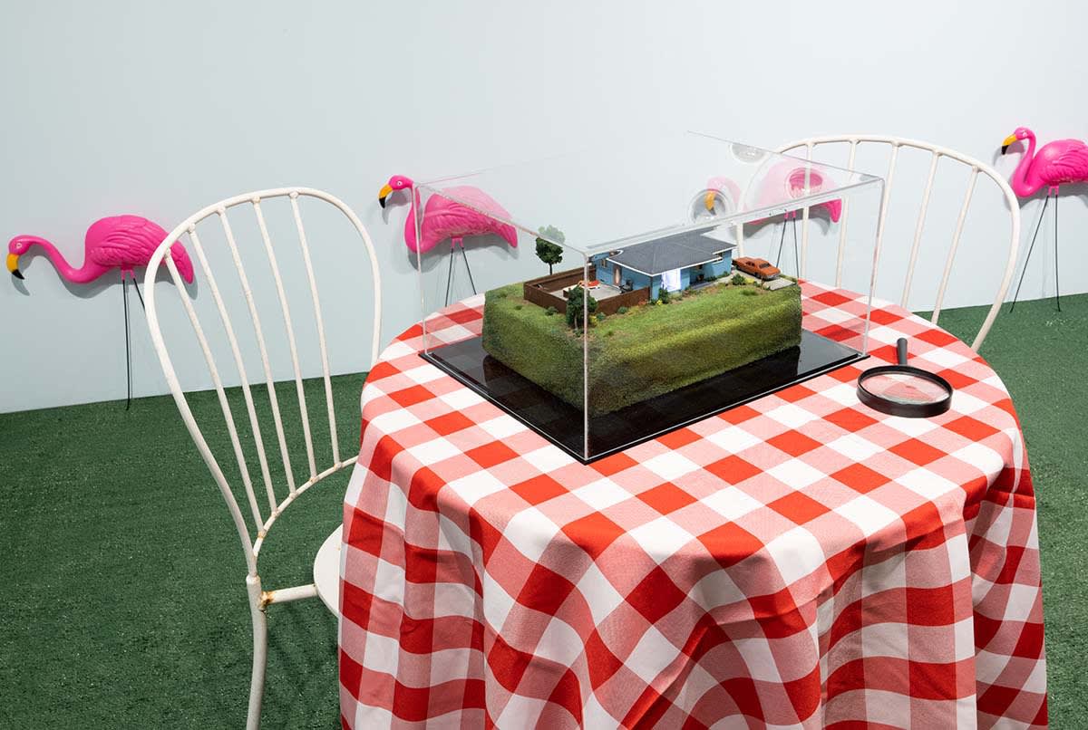 Abigail Goldman diorama sitting on red checkered tablecloth with magnifyer. Turf on ground, pink flamingos in background
