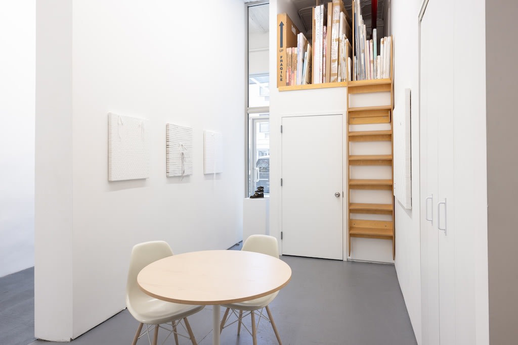 Installation of Didi Rojas exhibition - shoelace woven paintings on wall; round table with white chairs in foreground