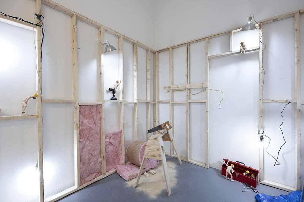 wooden scaffolding on white walls with pink insulation, a wooden work bench with saw, and dog person sculptures at Stephen Morrison Dog Show #4: House Broken exhibit