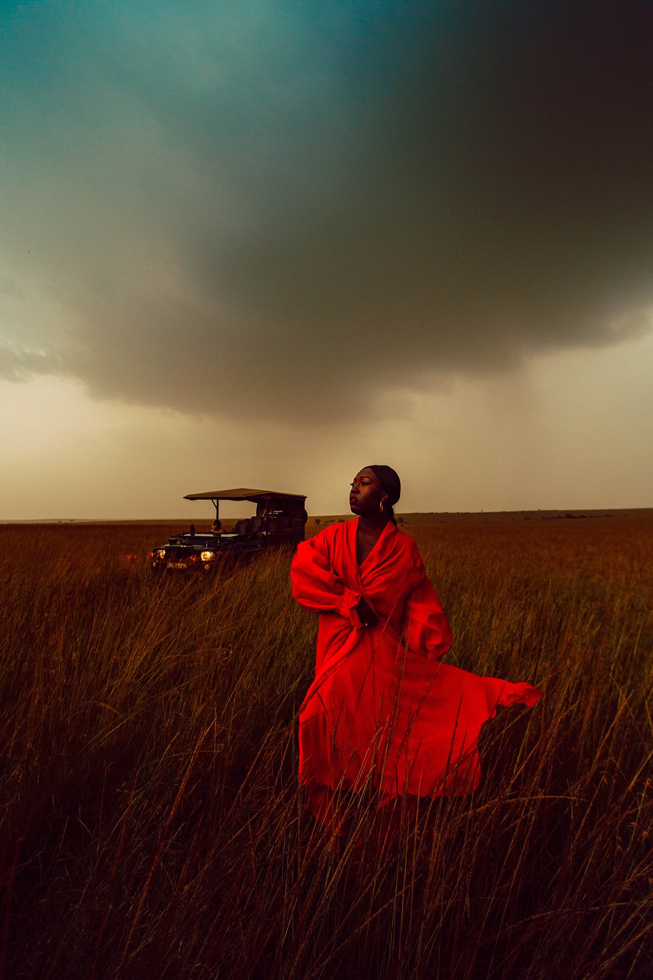 Black woman in scarlet red dress in the field in Tanzania, Africa, by Colby Blount, red dress floating in the wind, an old car in the background