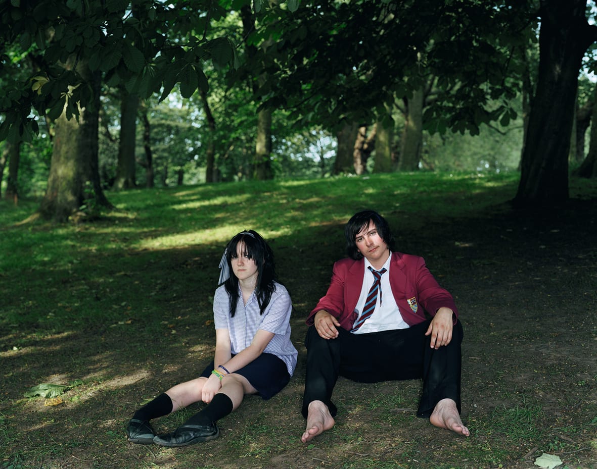 A Rineke Dijkstra photograph: 2 teenagers in school uniforms, one female and one male, sit under a tree on a grassy hill.