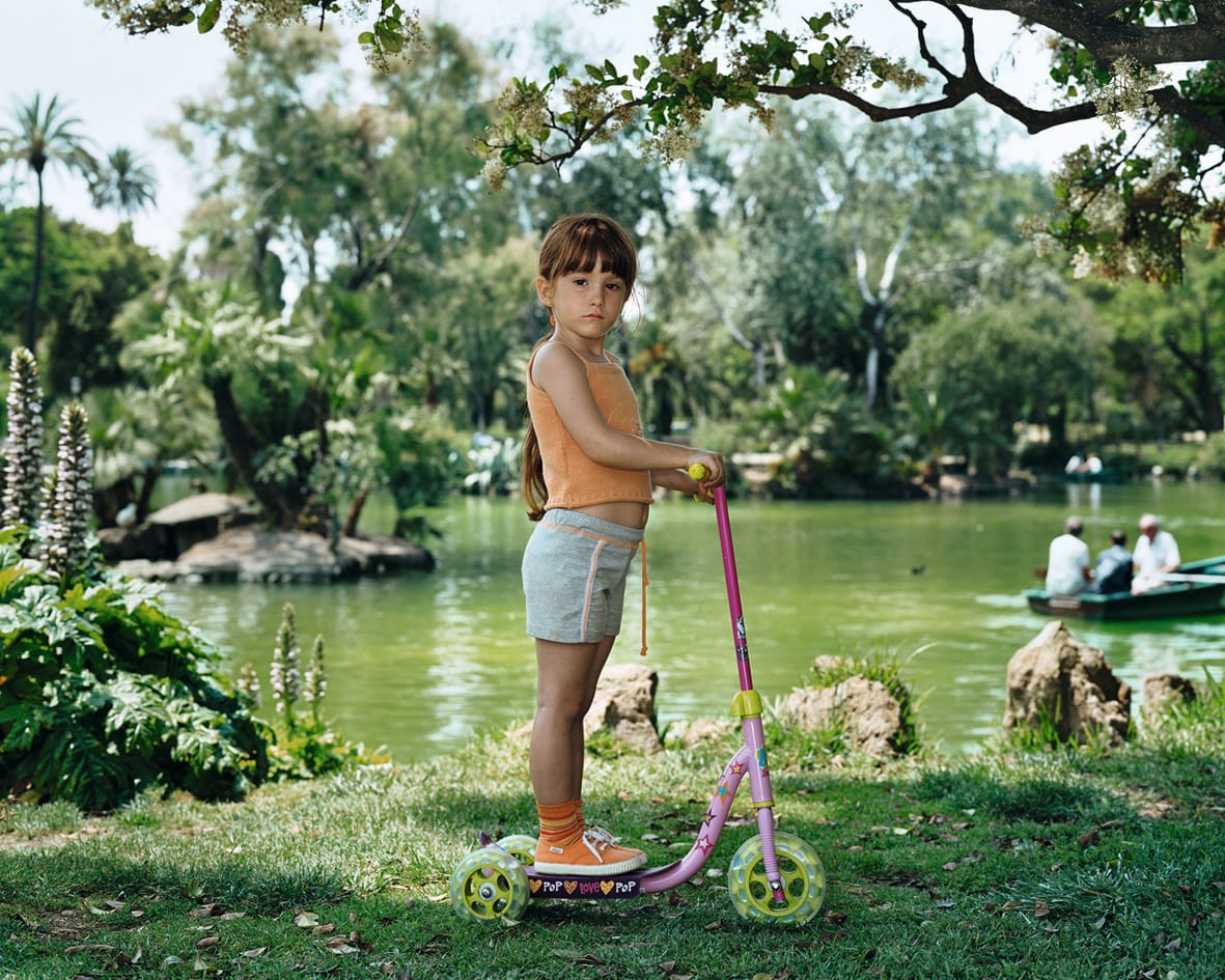 A Rineke Dijkstra photograph: A girl stands on a pink scooter in front of a green pond. Rowboats are out of focus in the background.