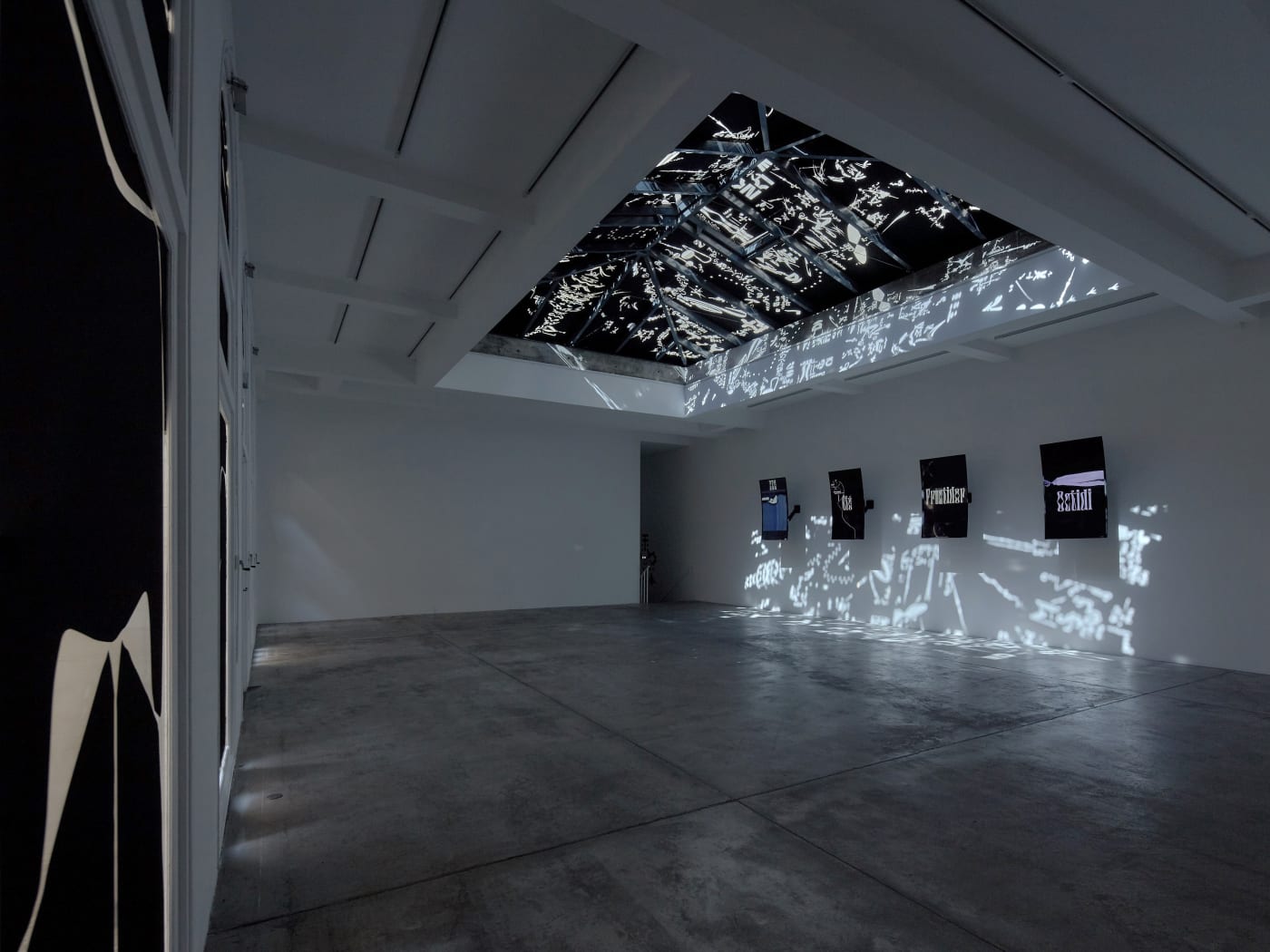 A wide installation view of Adrián Villar Rojas' Paris exhibition; light coming through decals over 4 LCD screens on wall