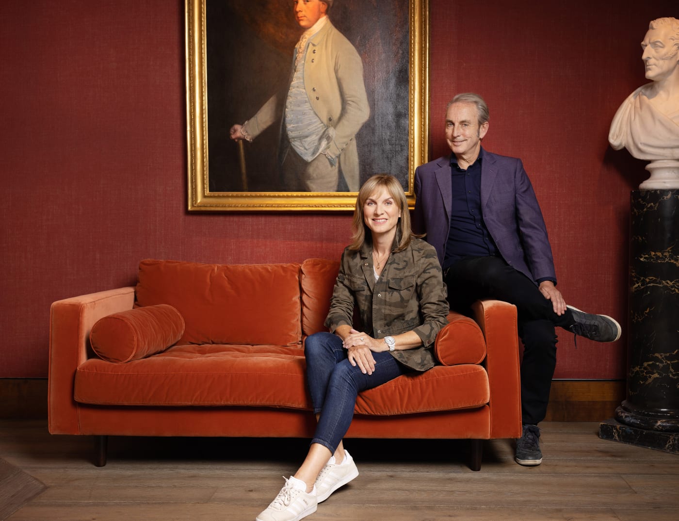 fiona bruce and philip mould sitting on a sofa in a red gallery with a painting