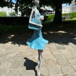 Fifth Avenue, zeitgenössische Frauenskulptur Hedwige Leroux, ein junges Mädchen in einem blauen Kleid, das im Wind weht, während sie einen kleinen Hund in ihrer Tasche trägt, Bronzeskulptur Art Yi-Galerie Brüsseler Kunstgalerie