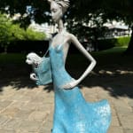 Fifth Avenue hedendaagse vrouwensculptuur Hedwige Leroux, een jong meisje onder een blauwe jurk die in de wind waait terwijl ze een kleine hond in haar tas draagt, bronzen sculptuur Art Yi-galerij Kunstgalerij Brussel