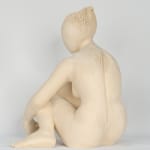 Seated white ceramic figure is looking over her left shoulder with arms draped around her knees which are tucked together.