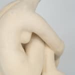 Seated white ceramic figure is looking over her left shoulder with arms draped around her knees which are tucked together.