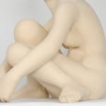 Seated white ceramic figure is looking over her left shoulder with arms draped around her knees which are tucked together.