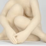 Seated white ceramic figure is looking over her left shoulder with arms draped around her knees which are tucked together.