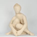 Seated white ceramic figure is looking over her left shoulder with arms draped around her knees which are tucked together.