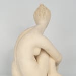 Seated white ceramic figure is looking over her left shoulder with arms draped around her knees which are tucked together.