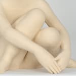Seated white ceramic figure is looking over her left shoulder with arms draped around her knees which are tucked together.