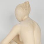 Seated white ceramic figure is looking over her left shoulder with arms draped around her knees which are tucked together.