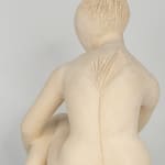 Seated white ceramic figure is looking over her left shoulder with arms draped around her knees which are tucked together.