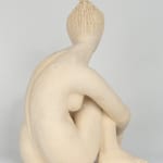 Seated white ceramic figure is looking over her left shoulder with arms draped around her knees which are tucked together.