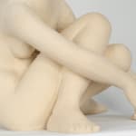 Seated white ceramic figure is looking over her left shoulder with arms draped around her knees which are tucked together.
