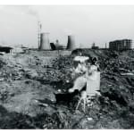 A fair maiden in white long skirt is putting on makeup and behind her and her dressing table, there are two industrial chimneys in the background