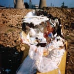 A fair maiden in white long skirt is putting on makeup and behind her and her dressing table, there are two industrial chimneys in the background