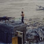 It is a video still from Farewell Poem 2002 that shows a worker standing on the top of roof is destroying the building near the river.