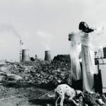 A fair maiden in white long skirt is putting on makeup and behind her and her dressing table, there are two industrial chimneys in the background