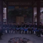 A video still from the section "Singing" of Chen Qiulin's work "Another Day" where shows a group of village women sitting around in a traditional wooden structure open-air building