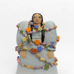 Ceramic female figurine modeled after ancient Assyrian votive figures. She wears a grey puffy dress and long black hair flows over her shoulders. She is wrapped in a colorful garland made of flowers and round, cylindrical, and canonical beads.