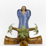 Ceramic female figurine modeled after ancient Assyrian votive figures. She stands in the center of four U-shaped palm trees that meet to hold her. Long black hair flows over her shoulders and she wears a V-neck, blue puffy dress, reminiscent of 1980s fashion.