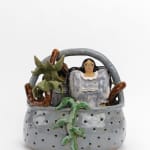 A ceramic grey basket with holes holds two small female figurines modeled after ancient Assyrain votive figures, palm trees, and leafy plants. The two figurines are back to back, dressed in puffy grey dresses with long black hair flowing over their shoulders.