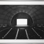 Framed black-and-white photograph of an empty theater with barrel-vaulted ceiling and an empty glowing white screen onstage