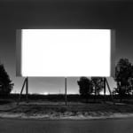 Black-and-white photograph of a blank glowing white screen and empty lot at night with city lights on the horizon
