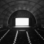 Black-and-white photograph of an empty theater with barrel-vaulted ceiling and an empty glowing white screen onstage