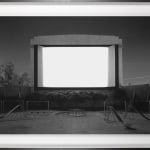 Framed back-and-white photograph of an empty glowing white screen set inside a bandshell standing above an empty playground at night