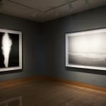 Lateral installation view of a framed vertical image of a splash of light next to a framed black-and-white photograph of a museum diorama of ice sheets floating on a calm sea