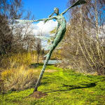 A monumental bronze sculpture of a winged female stands in front bushes and a lake at Dorset sculpture park, Sculpture by the Lakes