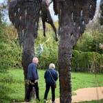 Two people are walking between the wings of the monumental bronze sculpture of a mythological figure at a Dorset sculpture park.