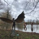 Bronze resin sculpture of mythical Greek winged figure Icarus by Nicola Godden, displayed on integral stone plinth, set in front of lake