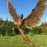 a bronze mythological winged figure, on grass in front of a hedge