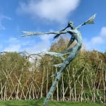 A monumental bronze sculpture of a winged female stands in front of a willow walk at a Dorset sculpture park, Sculpture by the Lakes