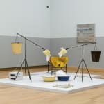 exhibition view of an installation consisting of two ceramic dog heads at the end of tripod stands. these figures are both balanced by buckets. the ceramic heads foam at the mouth, the foam then falls into large dishes which are fed back into the heads through pumps