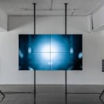 installation image of a large flat screen television suspended in air and sitting between two speakers on either side displays an image of two intersecting lines on a reflective surface.