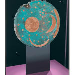 painted abstract circular image depicting sandy colored organic shapes atop a blue background, which is perched on an aluminum base