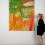 An image of a canvas painted in greens, reds, and oranges with the name Peter Stuyvesant painted on the right side. Next to the canvas, a figure wearing a black overcoat, skirt, and shoes gazes up at the piece.