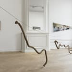 A sculpture of pieces of rope hanging from a string suspended over a wooden floor in a gallery.