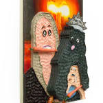 Mixed media hanging sculpture of a woman with a carrot in her mouth holding a small black dog with a Happy New Year paper crown on his head and a pretzel and a carrot in his hands. In the background an atomic bomb is going off.