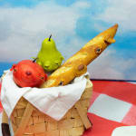 Installation featuring two bottles of wine with dog faces, two sandwiches with dog faces, a picnic basket with a baguette, an apple and a pear all with dog faces on top of a red and white checkered table clothe and a painted sky with clouds in the shape of dogs.
