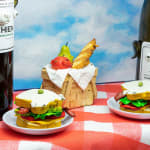 Installation featuring two bottles of wine with dog faces, two sandwiches with dog faces, a picnic basket with a baguette, an apple and a pear all with dog faces on top of a red and white checkered table clothe and a painted sky with clouds in the shape of dogs.