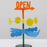 Metal sculpture with the word OPEN on the top and an arrow in orange. Stacked below are four yellow circles on yellow rods and stacked below those are blue berries and bananas on a steal stand.